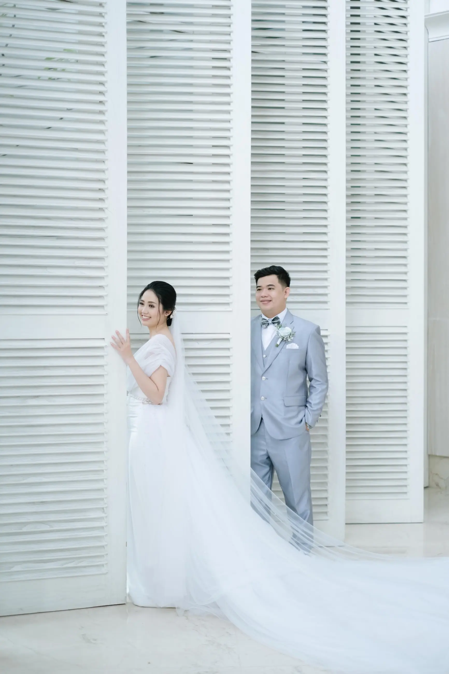 Pasangan pengantin berdiri di depan jendela krepyak putih, gaun dengan veil panjang